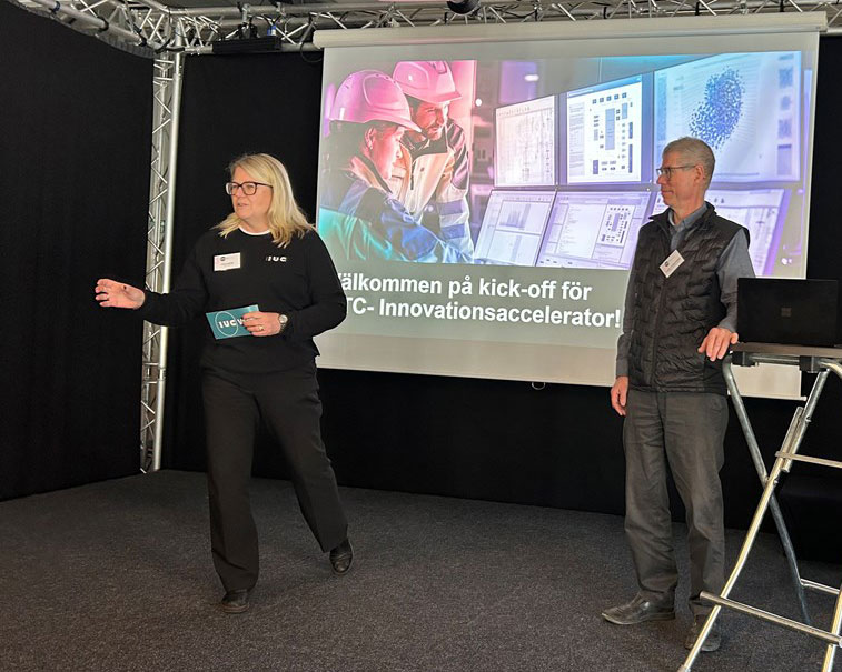 Två personer presenterar vid en kick-off för PTC Innovationsaccelerator, framför en stor skärm med bild på industrimiljö och datorer.