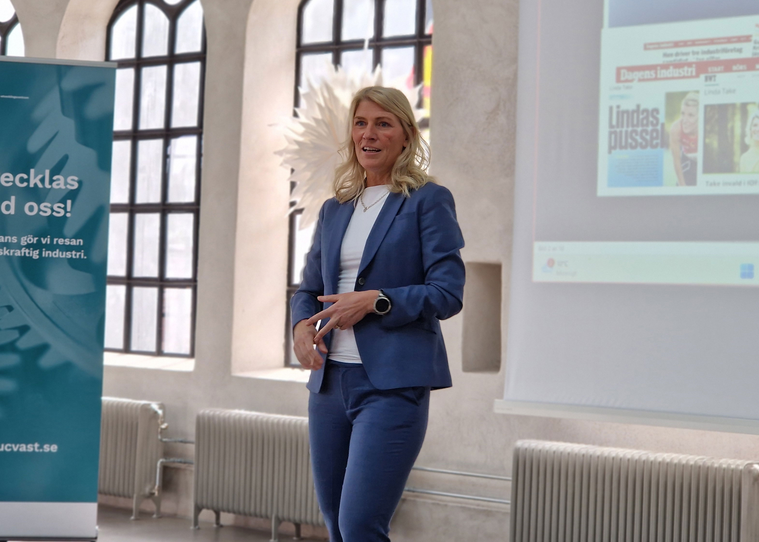 Föreläsare i blå kostym håller presentation framför duk i ljus konferenslokal.