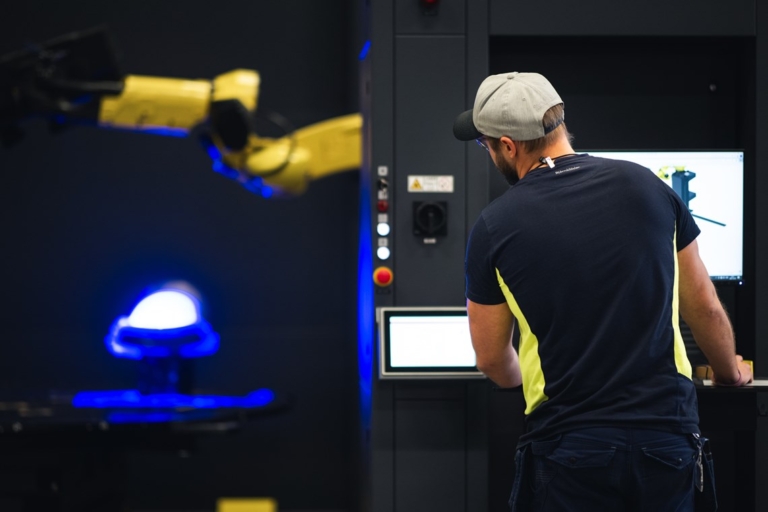 Person arbetar vid en datorstation och styr en gul robotarm i en avancerad industriell miljö.
