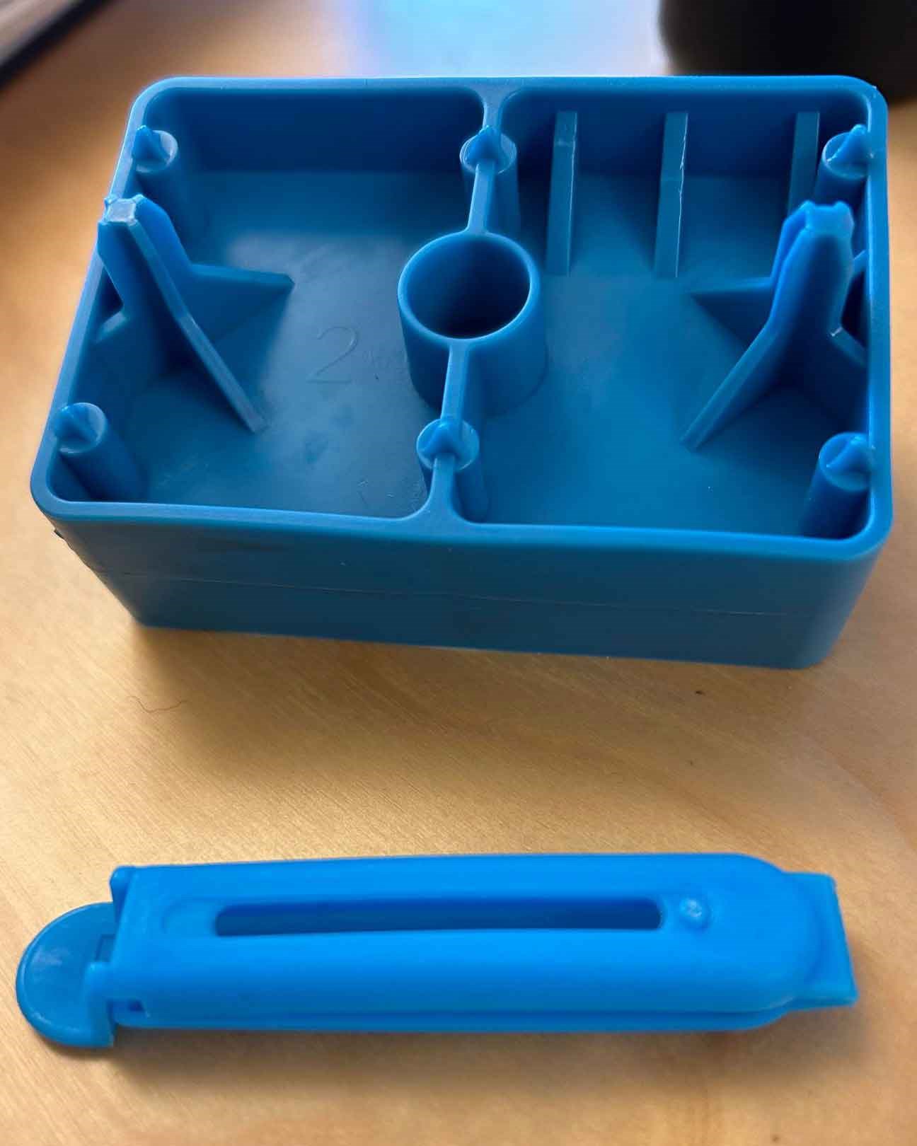 Två blå formsprutade plastdetaljer placerade på ett bord, en rektangulär låda med inre stöd och en långsmal del bredvid.