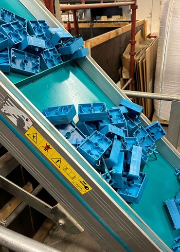 Blå formsprutade plastdetaljer rullar fram på ett grönt transportband i en industriell produktionsmiljö.
