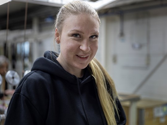 Ung kvinna med långt blont hår och svart hoodie ler i industrimiljö.