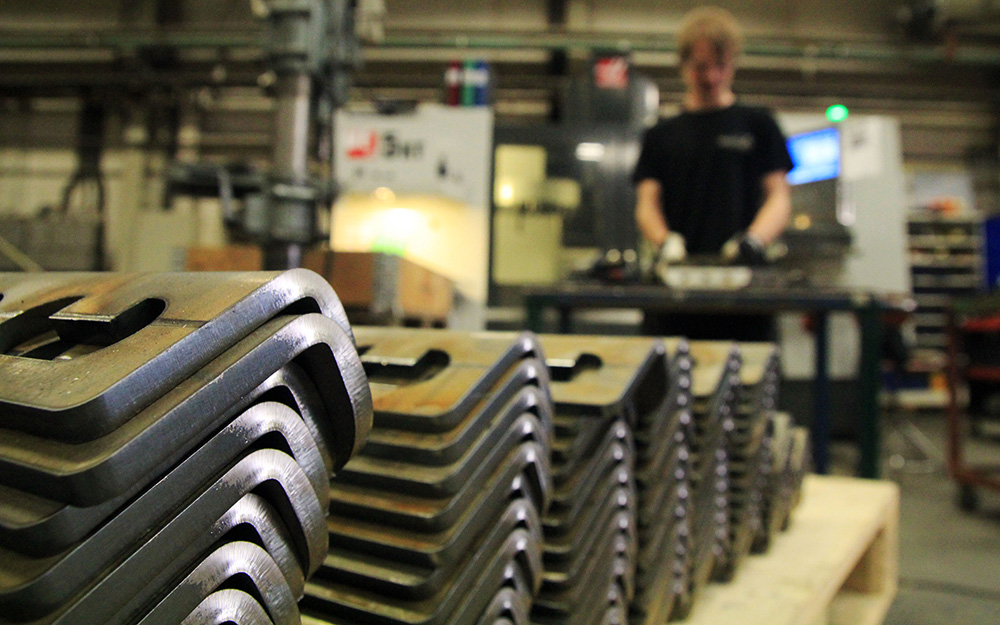 Staplar med bearbetade metallkomponenter i förgrunden, operatör i bakgrunden vid maskin.
