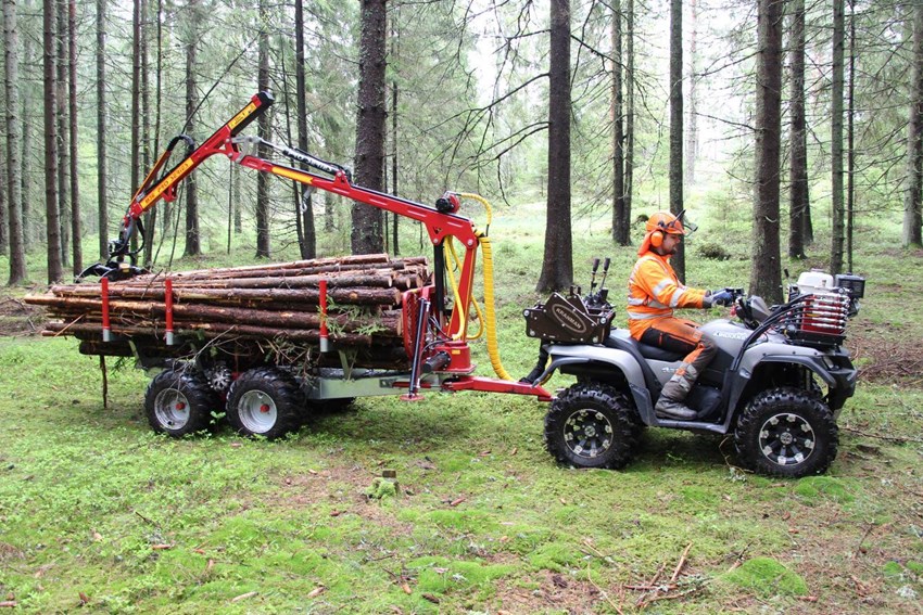 En person i orange skyddsutrustning använder en ATV med griplastarvagn från Kranman för att lasta timmer i en tät barrskog.