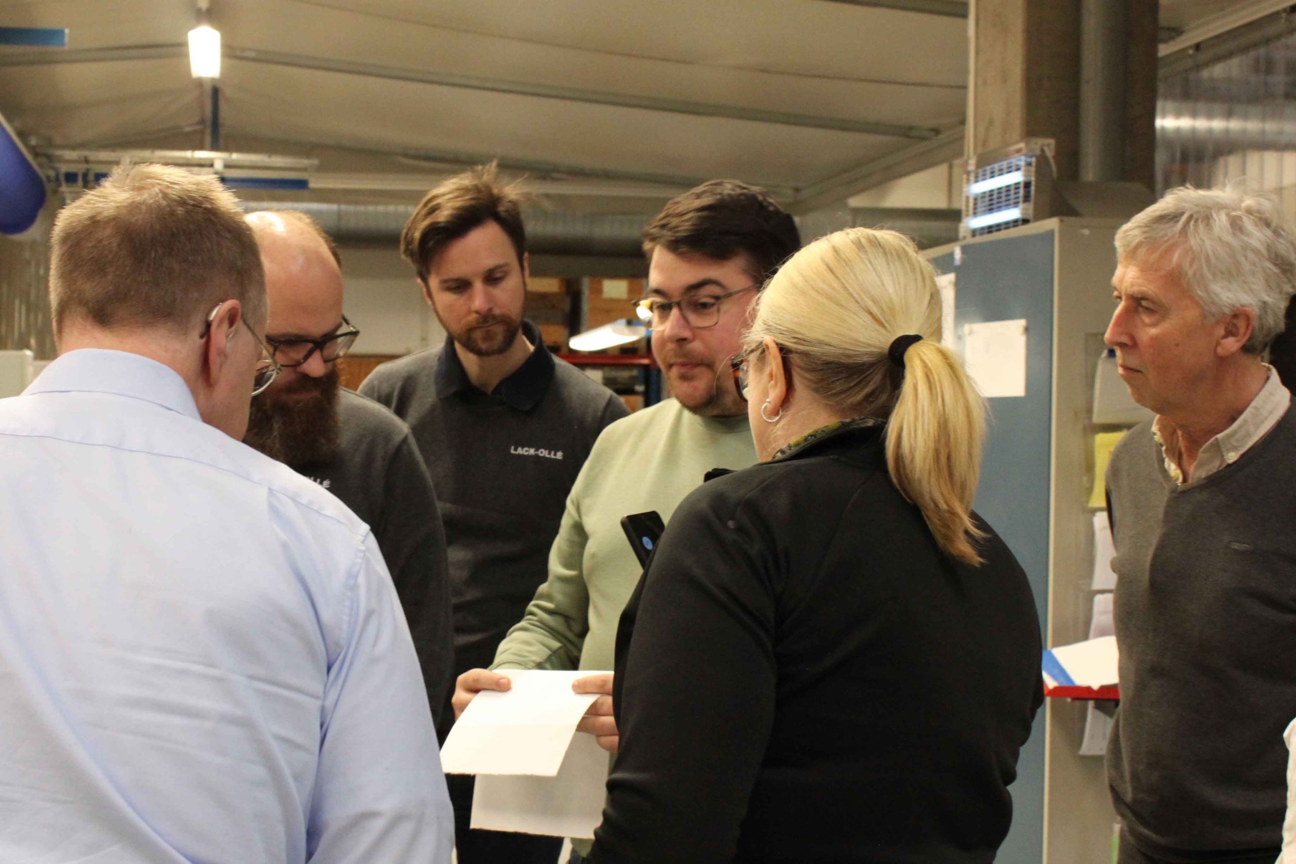 Grupp personer i arbetskläder samlas kring ett dokument i en industriell miljö.