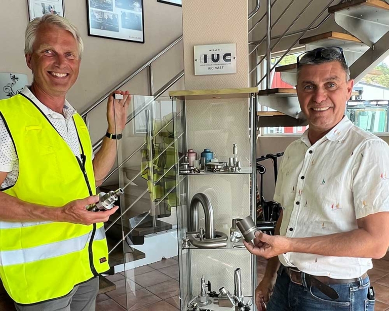 Två leende män visar upp metallkomponenter tillverkade med precision vid en monter i industriell miljö med IUC Väst-skylt i bakgrunden.