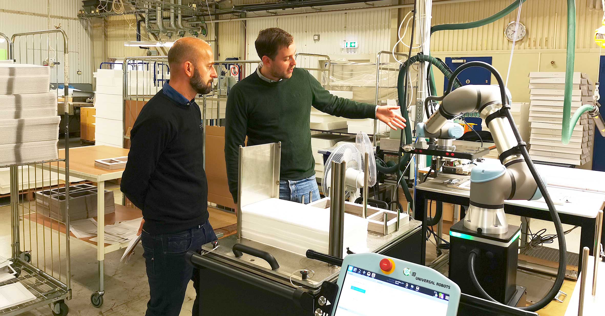 Två män diskuterar automationslösning med robotarm i en industriell miljö.