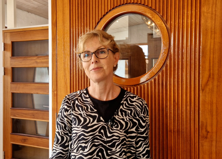 Leende kvinna med glasögon och zebramönstrad kavaj står framför en rund träpanel med fönster och pratar om företagets policy om hållbarhet.