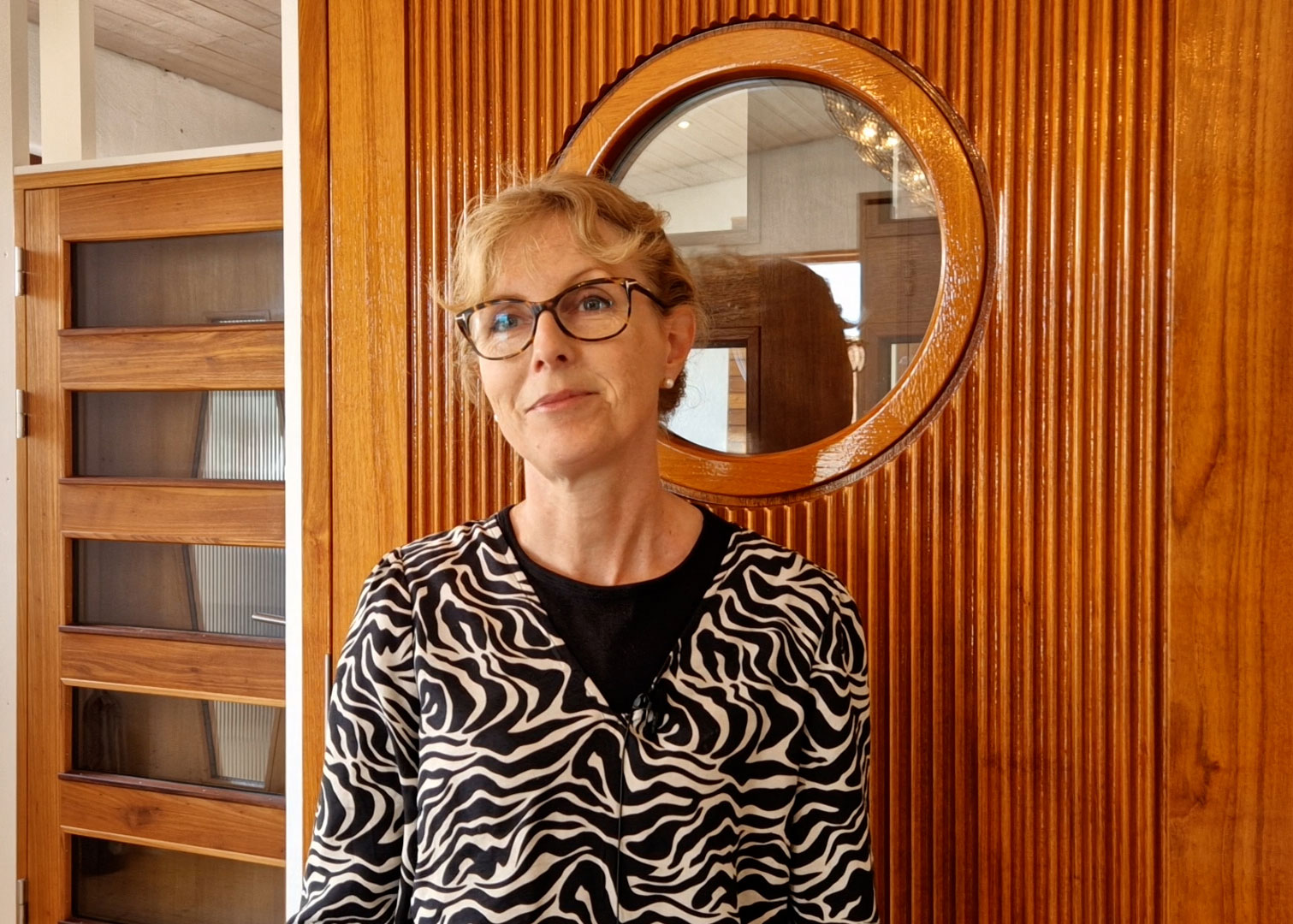 Leende kvinna med glasögon och zebramönstrad kavaj står framför en rund träpanel med fönster.