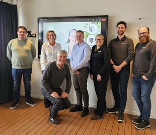 Arbetsgrupp poserar framför en presentationsskärm med digitalt mötesmaterial.