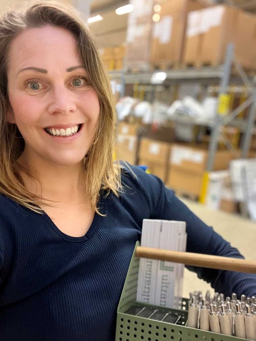 Leende kvinna håller i korg med tryckt material och pennor i lagerlokal.