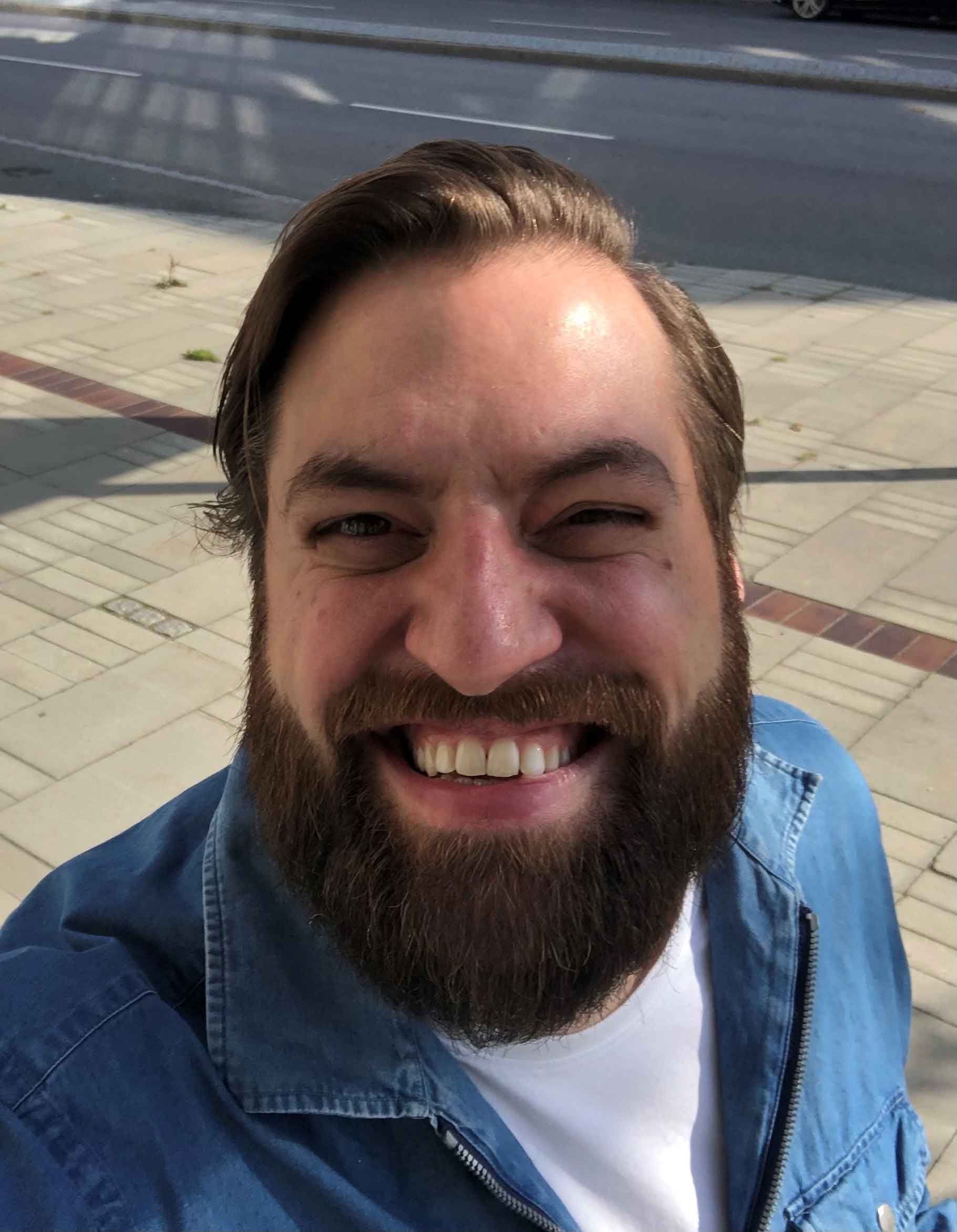Man med skägg ler stort i solljus, iförd blå skjorta utomhus