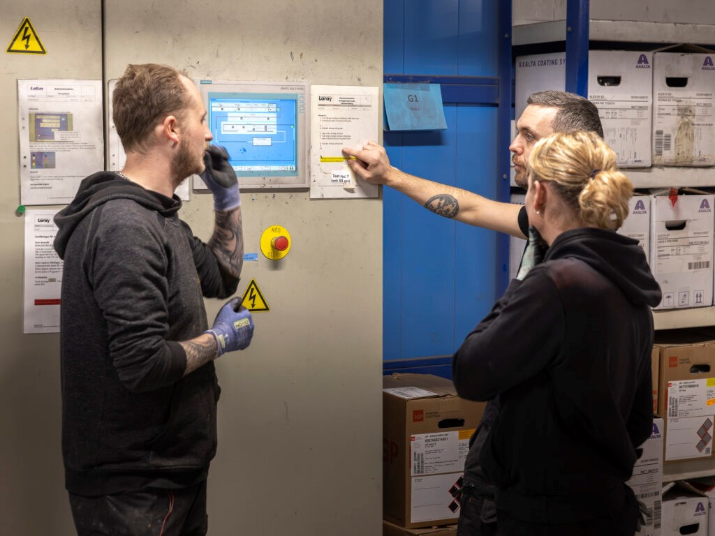Tre personer i arbetskläder diskuterar produktion vid en vägg med instruktioner.