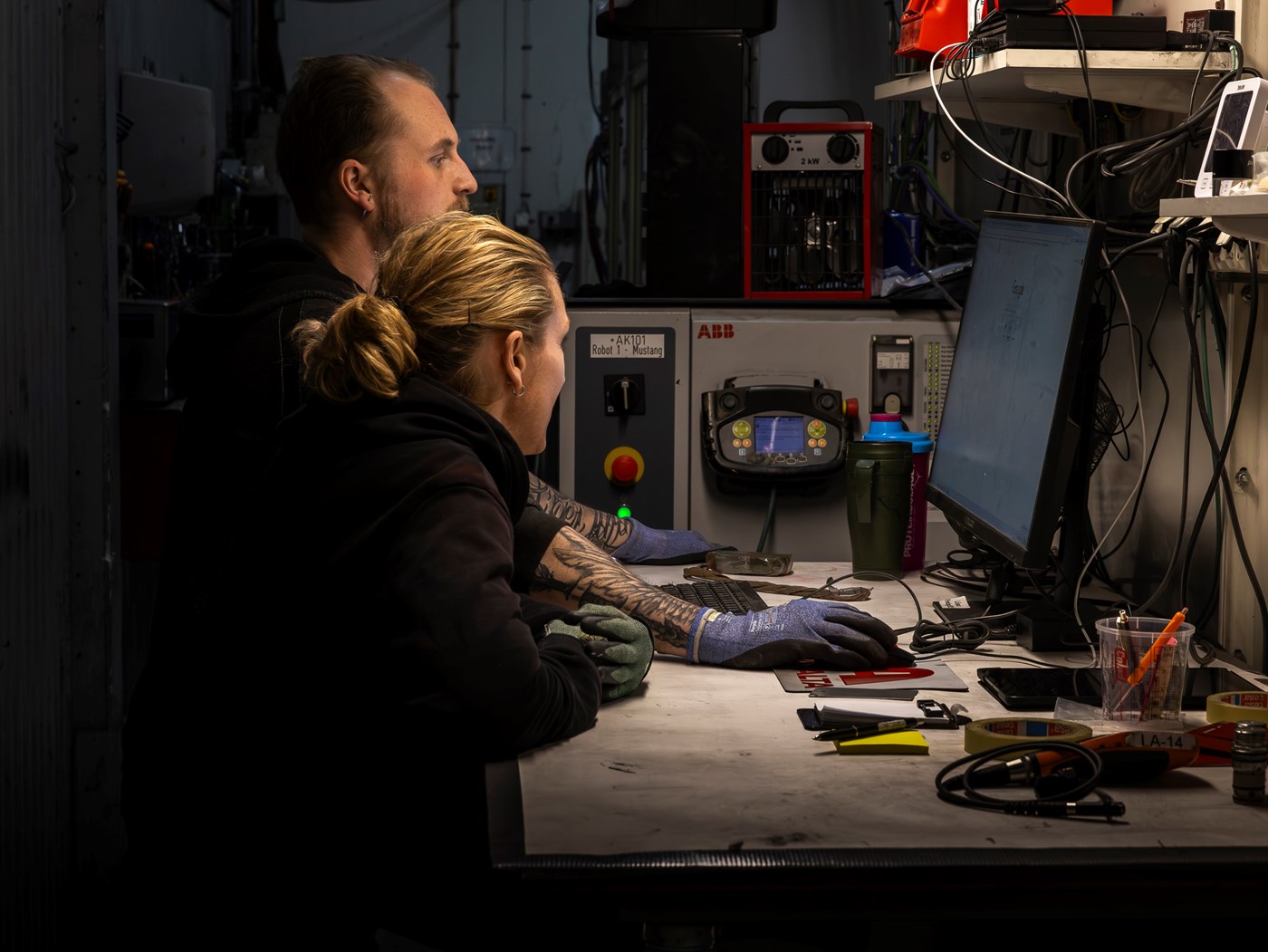 Två personer i arbetskläder arbetar vid dator i industrimiljö med teknisk utrustning.