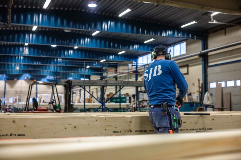 SJB-medarbetare i blå tröja arbetar med träkonstruktion i en stor industrilokal.