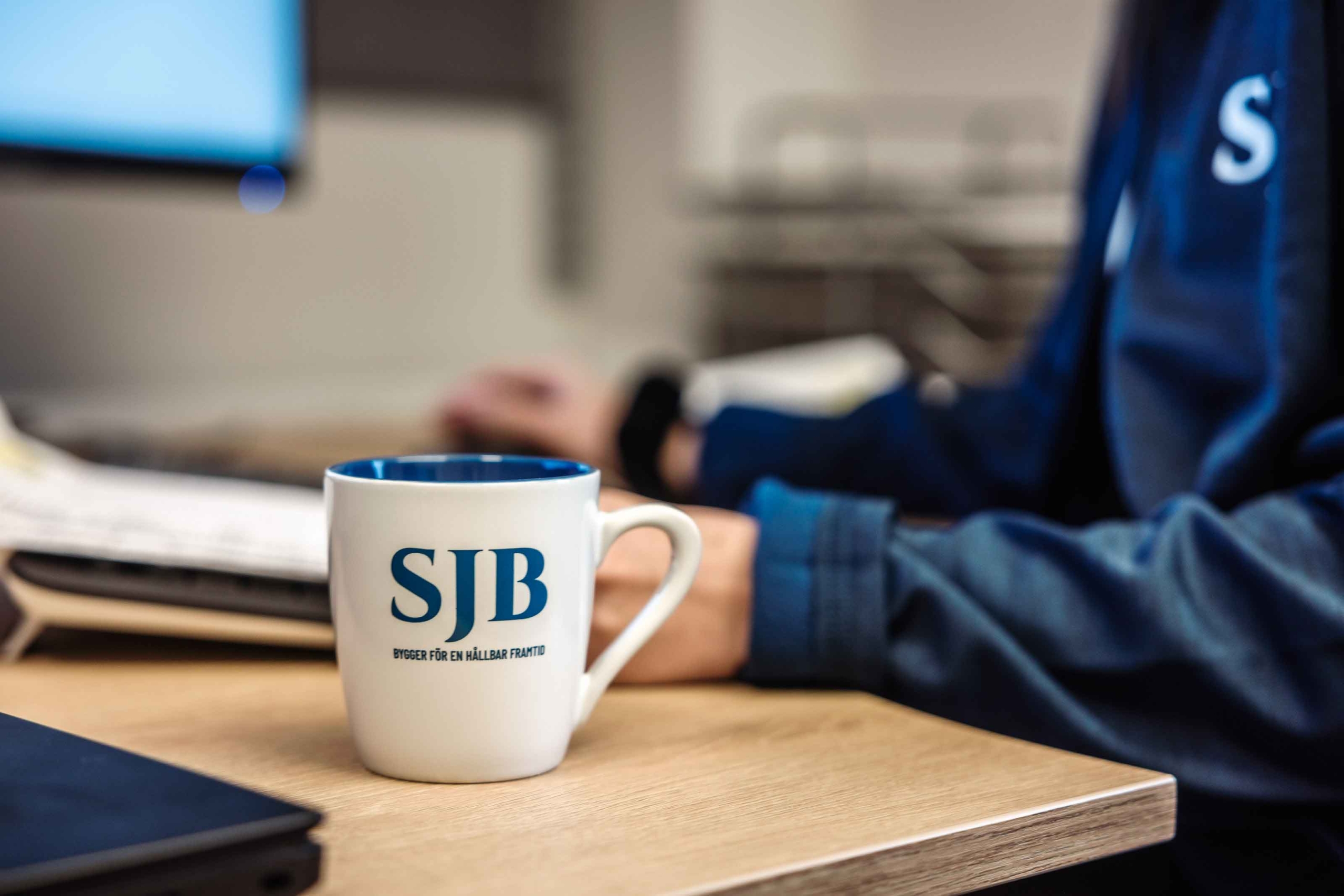 Närbild på en vit kaffekopp med SJB-logotyp och sloganen "Bygger för en hållbar framtid", på ett skrivbord.