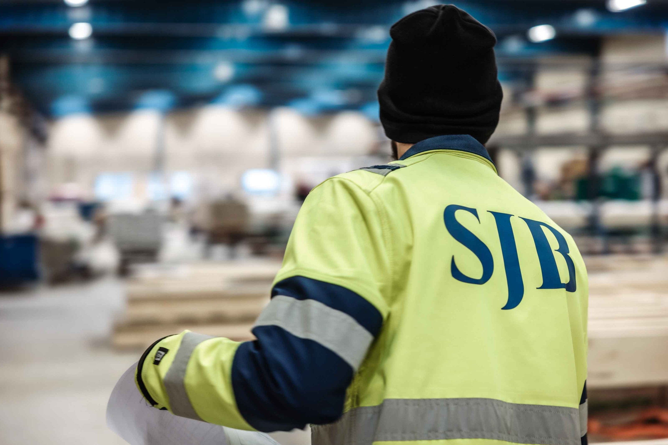 Person i gul SJB-arbetsjacka i fokus, stående i en industrihall med oskarp bakgrund.