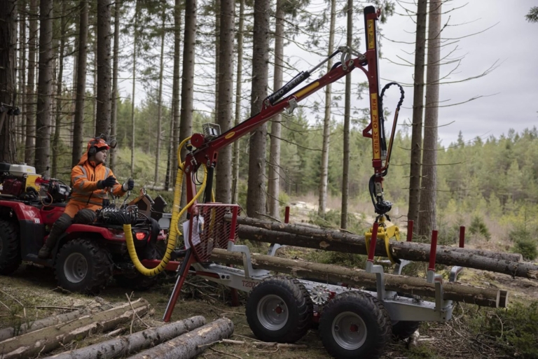 En griplastarvagn fullastad med timmer dras av en fyrhjuling genom en grön skog av gran och tall.