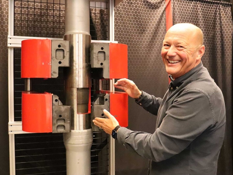 Tommy Tveter visar upp en industriell komponent med röd hållare och kraftiga metallrör i en teknisk testmiljö.