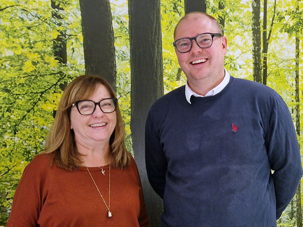 Två leende kollegor poserar framför bakgrund med grön skogsmotiv.