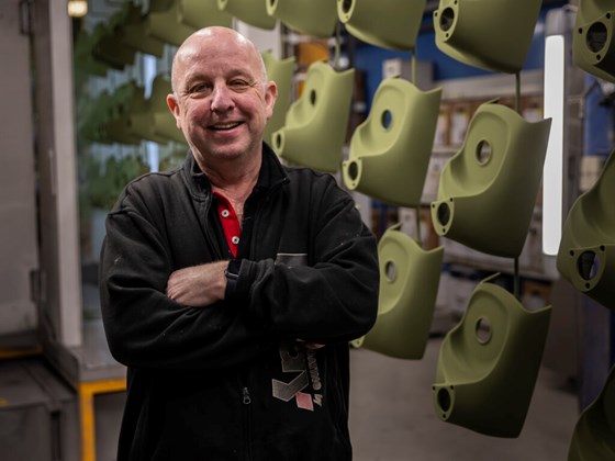 Leende man i svart jacka med armarna i kors framför upphängda industridetaljer.