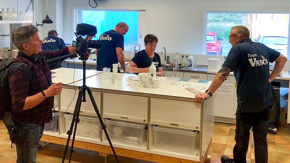 Filmteam dokumenterar laboratoriearbete hos Vasco, där personal i Team Vasco-tröjor arbetar vid labbbänk.