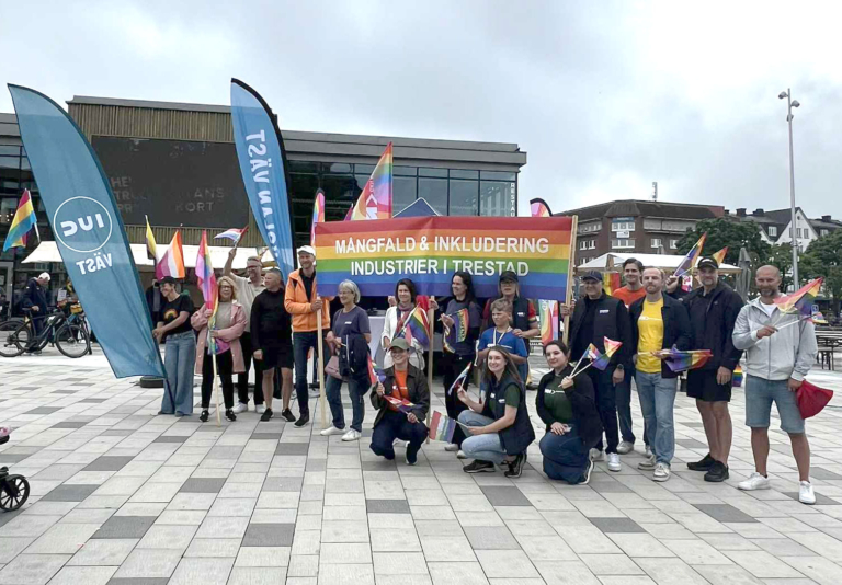 Gruppbild med deltagare som håller regnbågsflaggor och en banderoll med texten ”Mångfald & inkludering – industrier i Trestad” under Pride.
