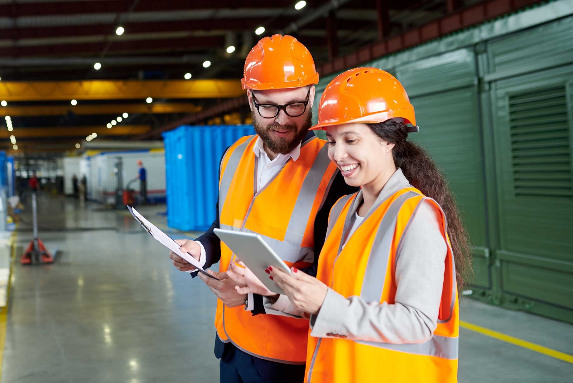 Man och kvinna i reflexvästar och hjälmar tittar på en surfplatta och ler i en industrihall. Handledarutbildning genom Teknikcollege.