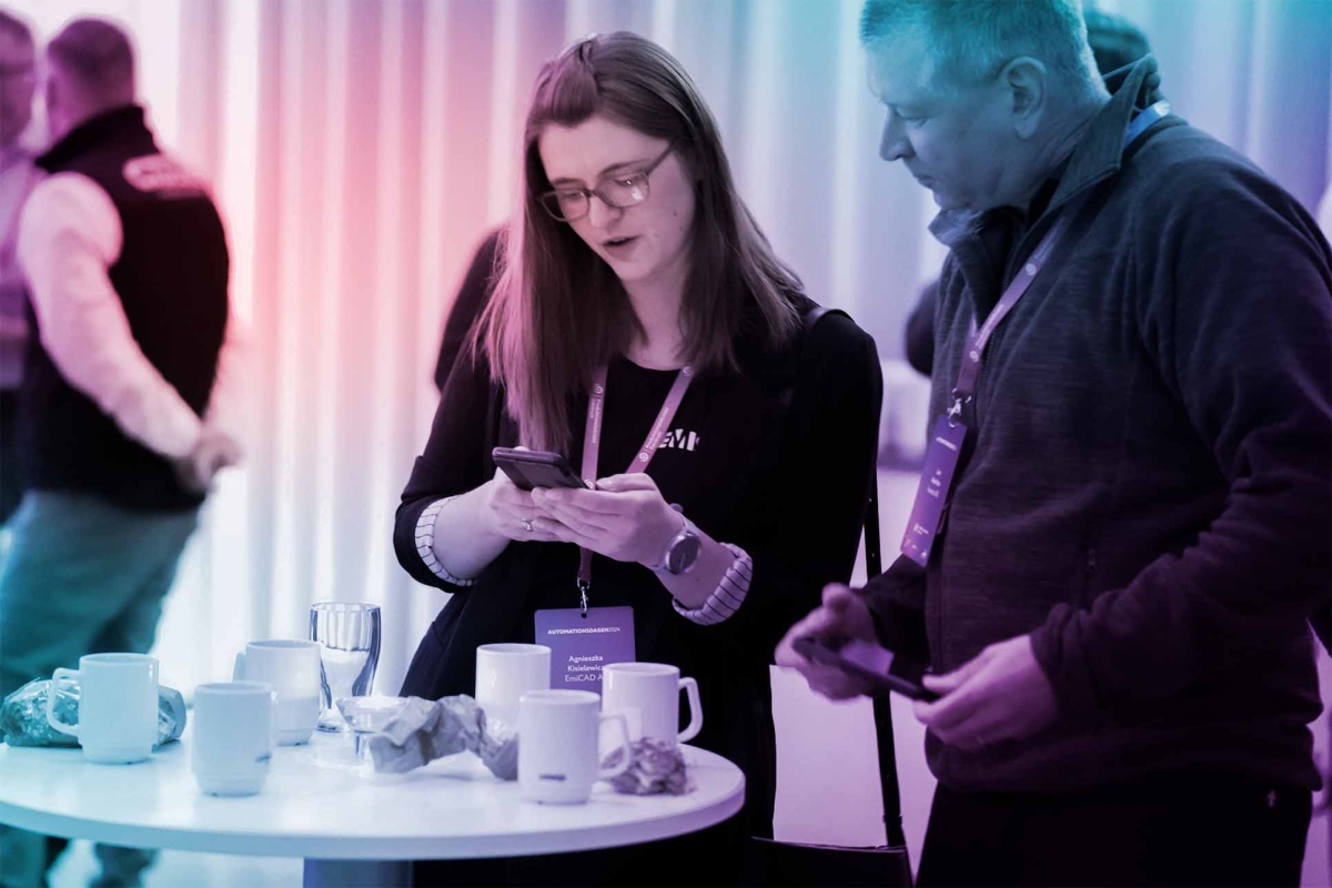 Två personer står vid ett bord under Automationsdagen och tittar på en mobil, omgivna av kaffekoppar och samtalande deltagare i bakgrunden.