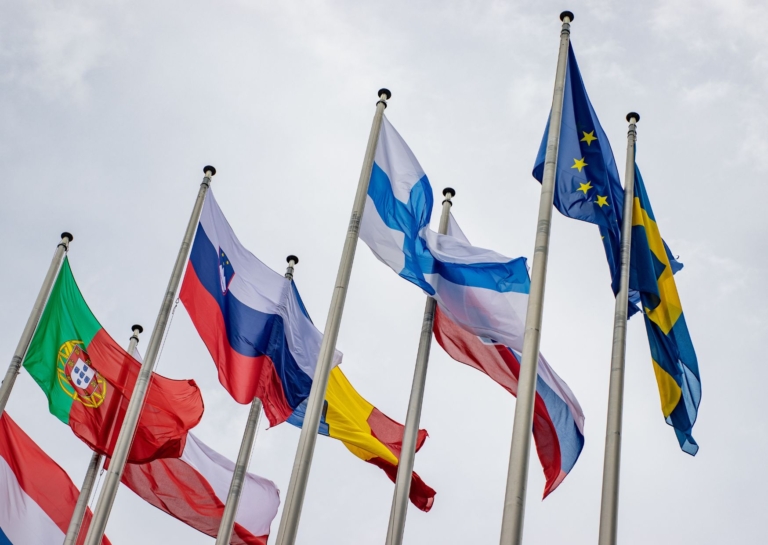 Flera EU- och medlemsstatsflaggor (bl.a. EU, Sverige och Finland) vajar på höga flaggstänger mot en molnig himmel.