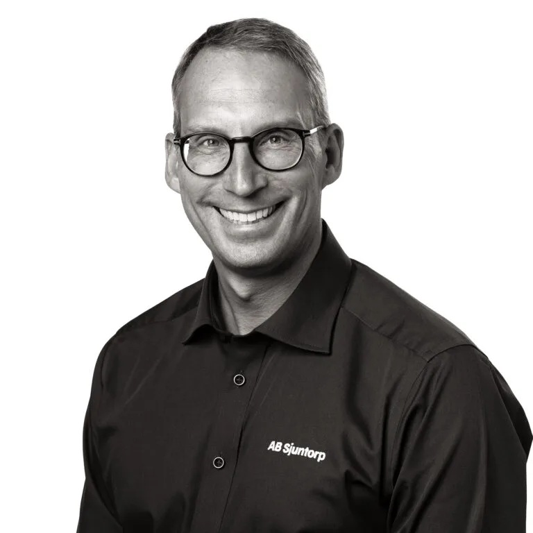 Porträtt av en leende man med runda glasögon i svart skjorta med logotypen ”AB Sjuntorp”, mot vit bakgrund.