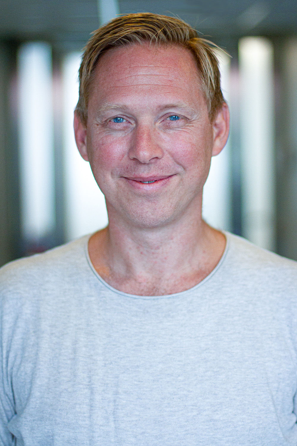 Porträtt av en leende blond man i ljusgrå tröja, mot en suddig inomhusbakgrund.
