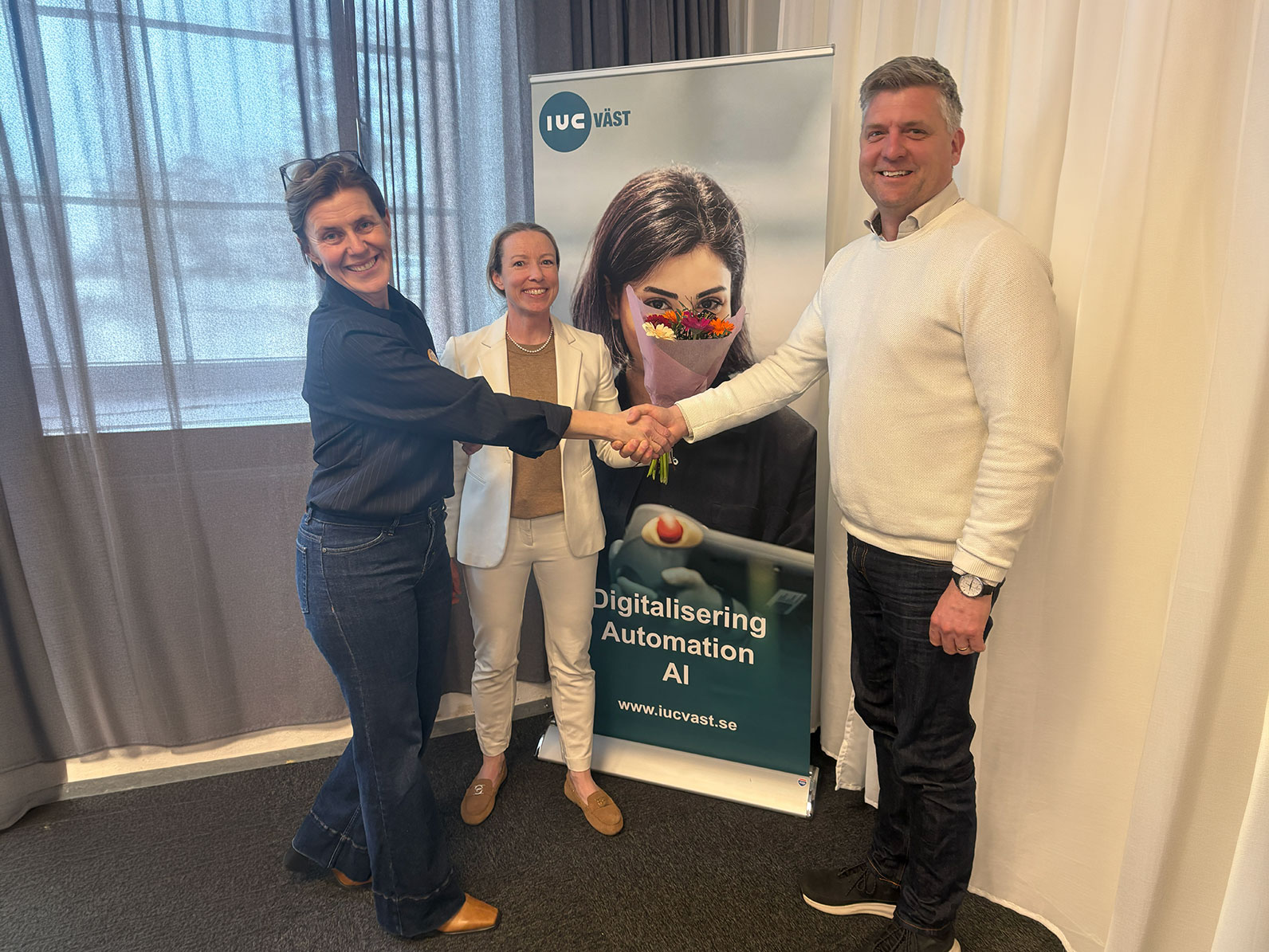 Tre personer står framför en rollup från IUC Väst och skakar hand under årsmötet.