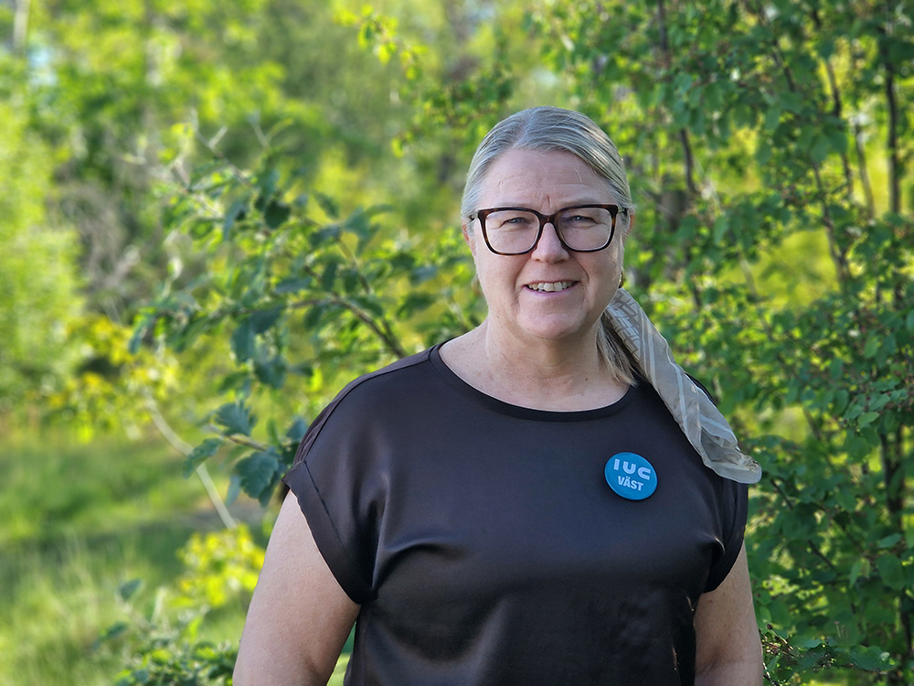 Lena André Olsson från IUC Väst står utomhus framför grönska. Hon arbetar med effektiv produktion inom Industry In West och hjälper industriföretag att utveckla arbetssätt, minska slöserier och stärka sin konkurrenskraft.